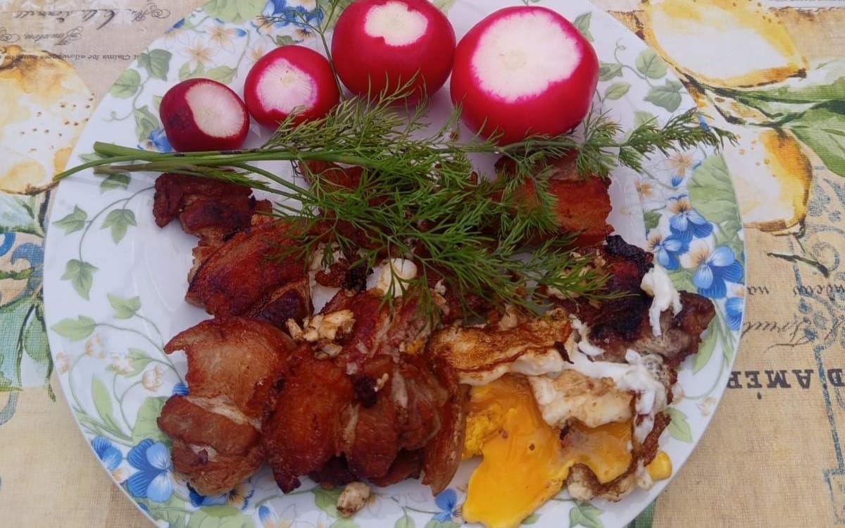 Тарелка деревенского завтрака с жареным мясом, яйцом и зеленью, рядом редис на скатерти