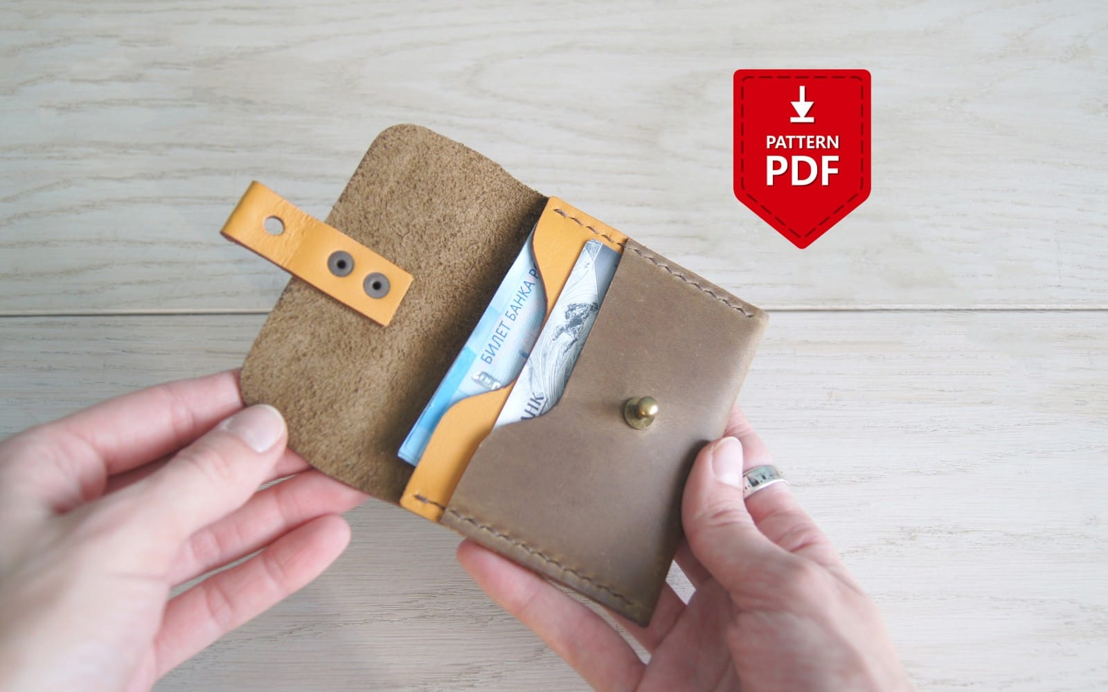 Hands holding a handmade leather fold card wallet with pattern PDF download badge on a wooden table